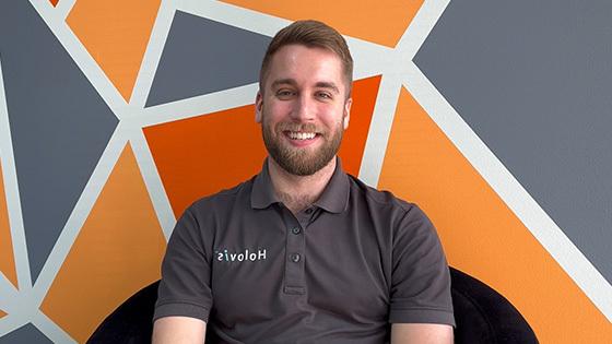 A man with light hair and a beard wearing a grey Holovis polo while seated and smiling in front of an orange, grey, and white geometric background.